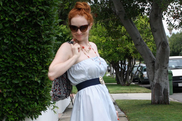 Woman with red hair in a light-blue strapless dress posing on a sidewalk beside hedges and trees, wearing sunglasses and a patterned backpack.