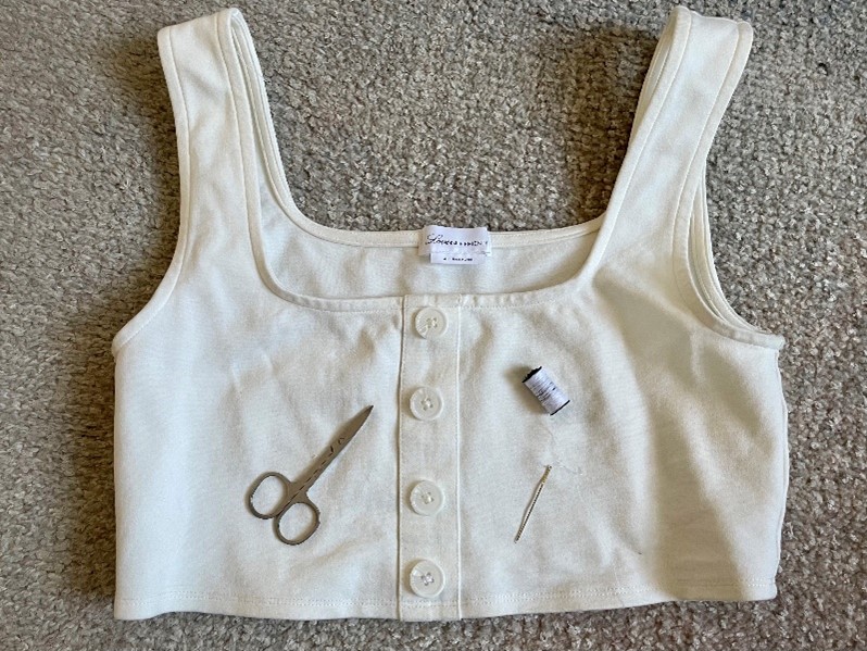 White cropped tank top with front buttons laid on carpet, with small scissors, thread, and a needle nearby.