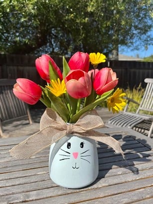 Whimsical blue bunny-face vase holds pink tulips and yellow daisy-like flowers on a sunlit wooden table.