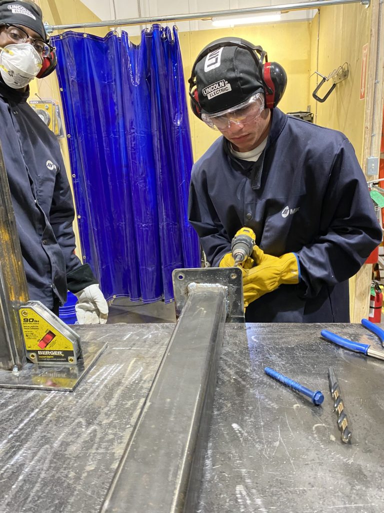 Two welders in navy coveralls and Lincoln Electric helmets grind a metal bracket at a workstation, blue curtain behind.