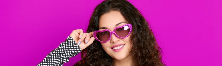 Woman with curly brown hair wearing glittery pink heart-shaped sunglasses against a bright pink background