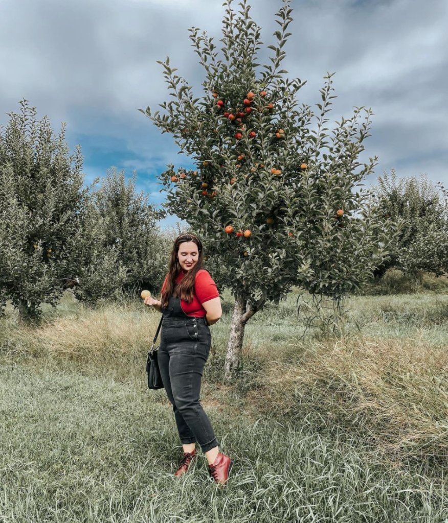 Woman in red shirt and black overalls posing beside an apple tree in a lush orchard, holding an apple.