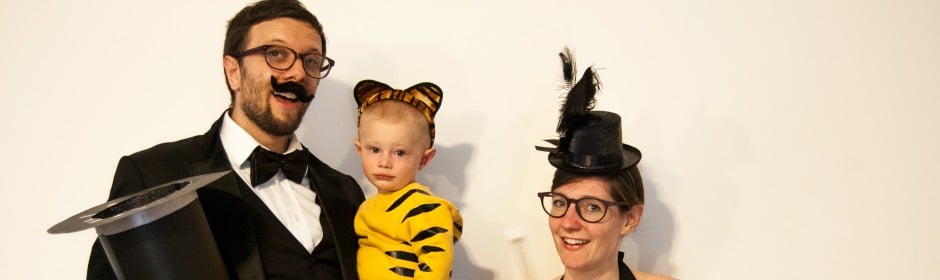 Three people pose in costumes against a white backdrop, wearing top hats, a tiger onesie, and feathered accessories.