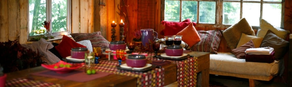 Cozy cabin dining nook with a wooden table set for a meal, candles, and colorful cushions along a windowed wall.