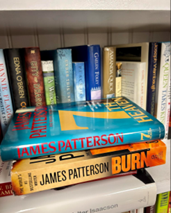 Stack of paperback thriller novels on a white shelf, dominated by James Patterson titles.