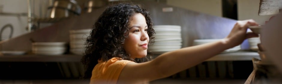 Woman with curly hair in an orange shirt reaching for plates in a busy kitchen.
