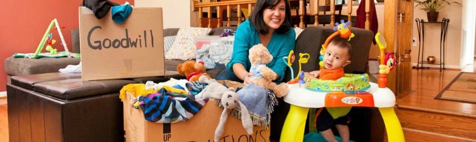 Mother and child with a Goodwill donation box among donated toys and blankets in a living room.