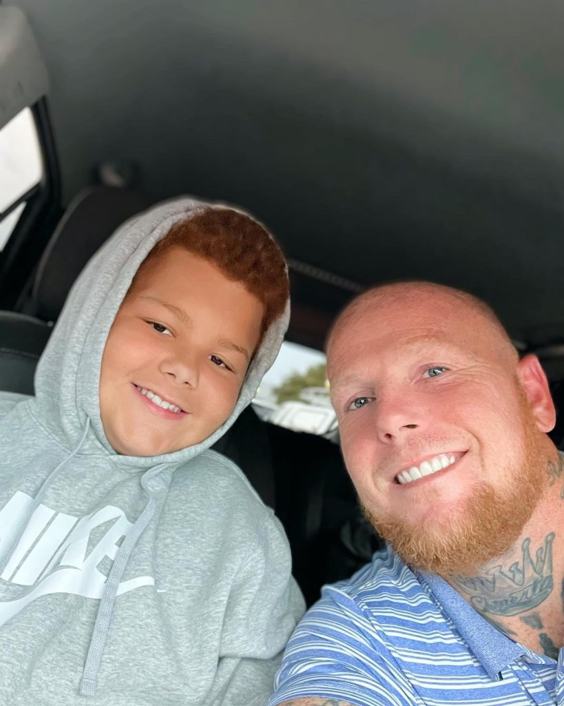 Smiling man with a beard and a boy in a gray hoodie take a selfie inside a car.