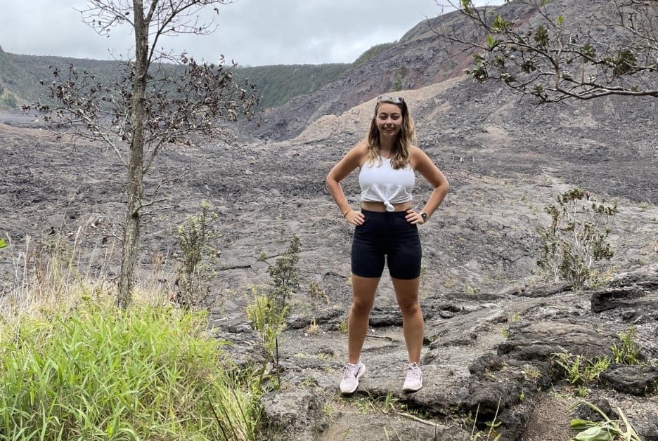 young woman standing on rocky volcanic landscape with sparse vegetation, wearing white crop top, black shorts and sneakers