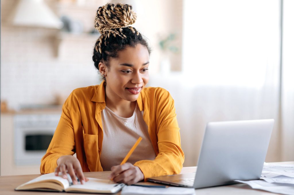 Young woman with a braided bun wearing an orange shirt, writing in a notebook beside a laptop.