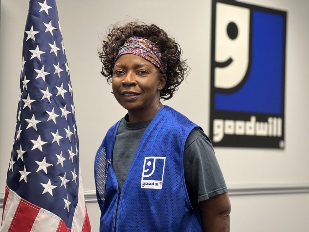 Black woman in a blue Goodwill vest stands beside the American flag inside a Goodwill facility.