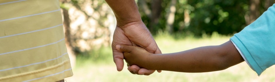 Two people hold hands outdoors in sunlight, one adult and one child.