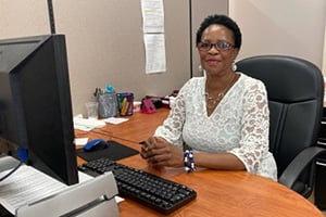 Office worker seated at a desk with a computer, wearing a white lace blouse and glasses.