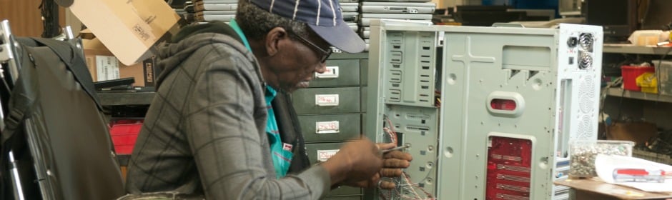 Technician in a teal apron and cap repairing a desktop computer inside a cluttered workshop.