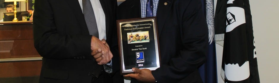 Two men in suits shake hands as one presents a framed award plaque with photos at a ceremony.