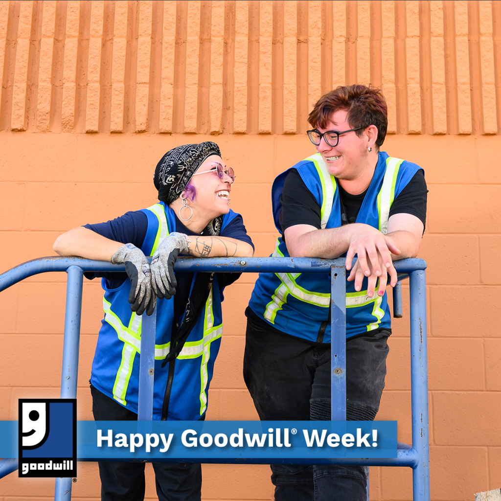 Two Goodwill workers in reflective vests lean on a blue railing, smiling at each other in front of an orange brick wall.