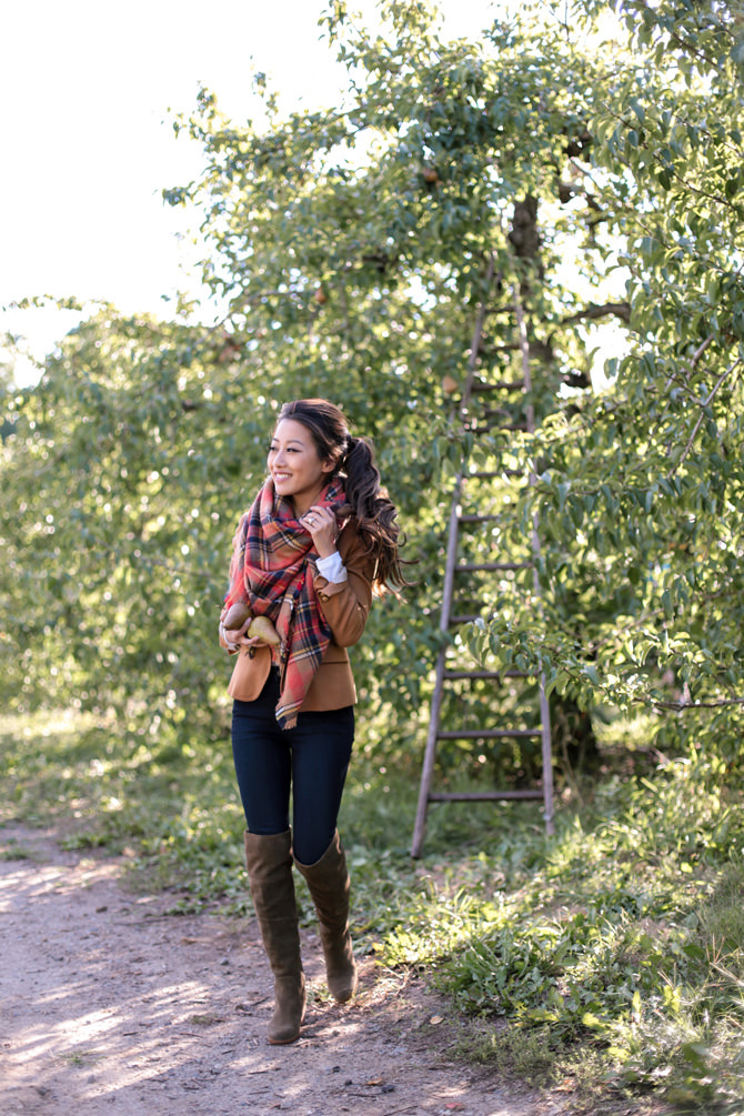 Smiling woman walking along a dirt path in an orchard, wearing a brown jacket, plaid scarf, and tall boots.