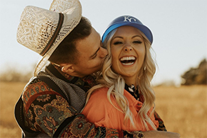 Couple embracing in a sunlit field; man kisses woman on the cheek as she smiles, wearing a blue cap.