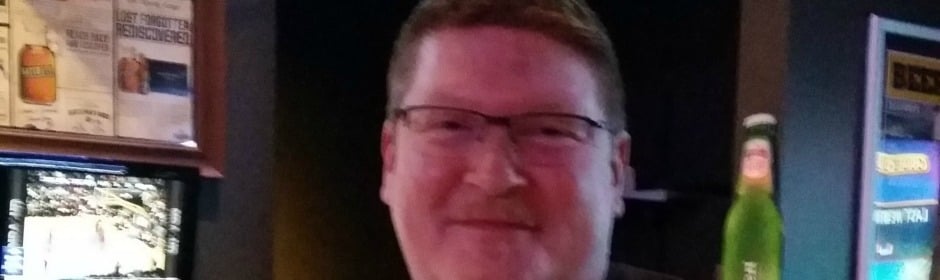 Smiling man with glasses in a dim bar, with a green beer bottle beside him.