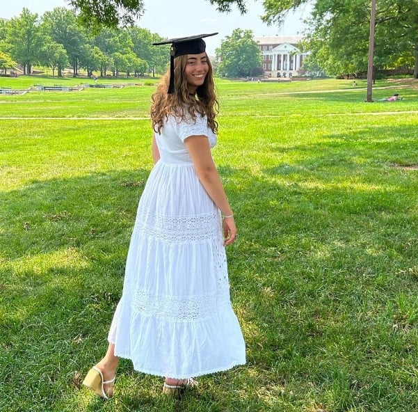 Graduate woman in a white dress and mortarboard standing on a sunny campus lawn, smiling at the camera.