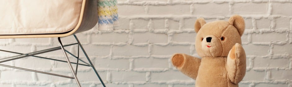 Beige upholstered chair with metal legs on the left and a tan teddy bear by a white brick wall.