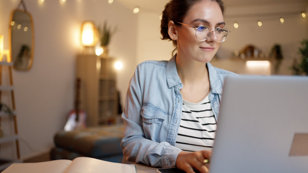 Young woman wearing glasses uses laptop in a warmly lit, cozy room.