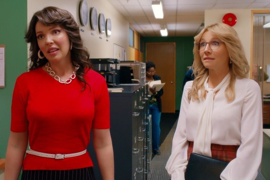 Two women in an office hallway: one in a red top with a white belt, the other in a white blouse holding a folder.