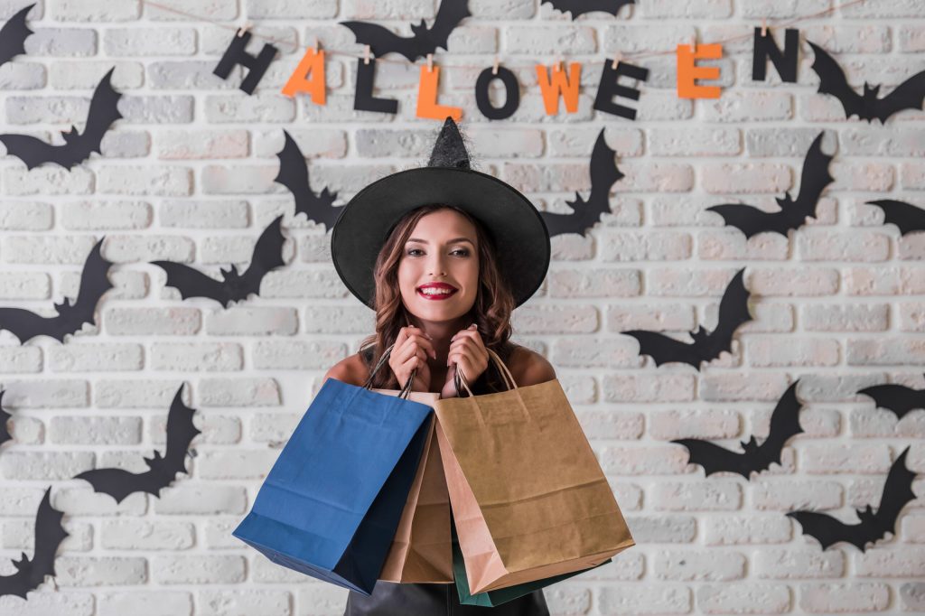 Woman wearing a witch hat holds blue and brown shopping bags in a Halloween backdrop with bat silhouettes and an orange HALLOWEEN banner.