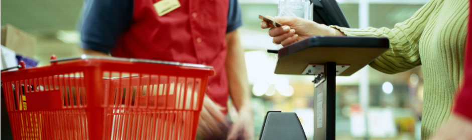 Shoppers at a supermarket checkout; red shopping cart in the foreground as a cashier scans items.