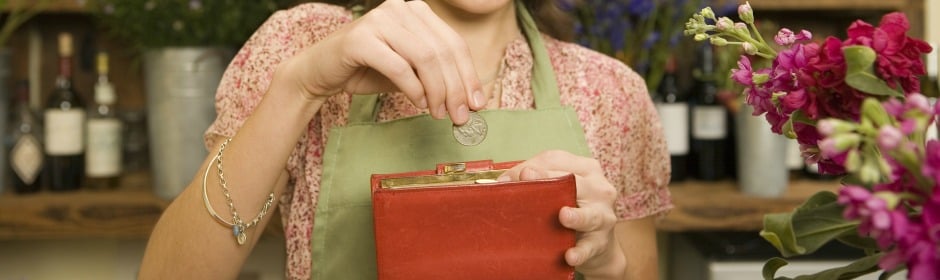 Person in a floral blouse and green apron places a coin into a red wallet at a shop counter, with flowers nearby.