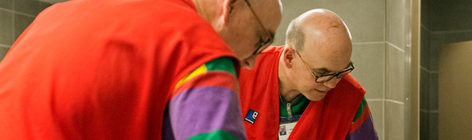 Two workers in red vests lean over a workstation, focused on a task in a tiled room.