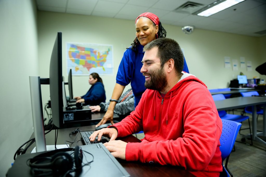 Man in a red hoodie types at a desktop computer while a female instructor in blue assists in a computer lab.