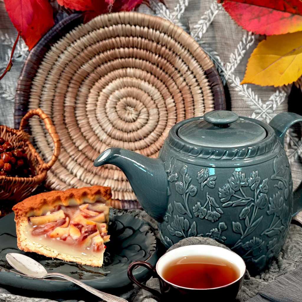 Teal ceramic teapot with floral relief beside a slice of fruit tart and a cup of tea on a gray table, surrounded by autumn leaves.