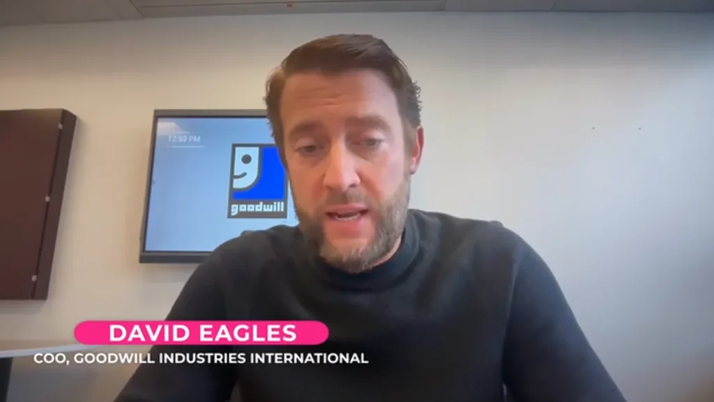 Man in a dark sweater speaks in a video call with a Goodwill logo on a monitor behind him.