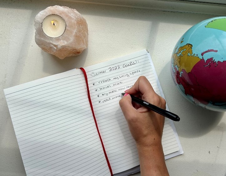 Hand writing in a lined notebook titled Summer 2023 Goals, with a candle and globe nearby.