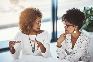 Two women chat at a bright table, one gesturing with hands as the other holds a cup.