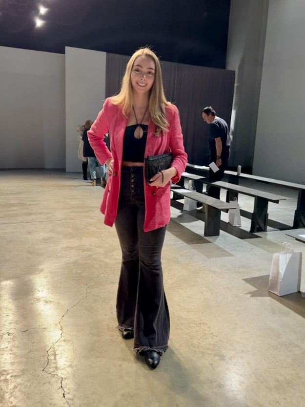Woman in a pink blazer, black top, and dark bell-bottoms posing in a spacious hall with benches.