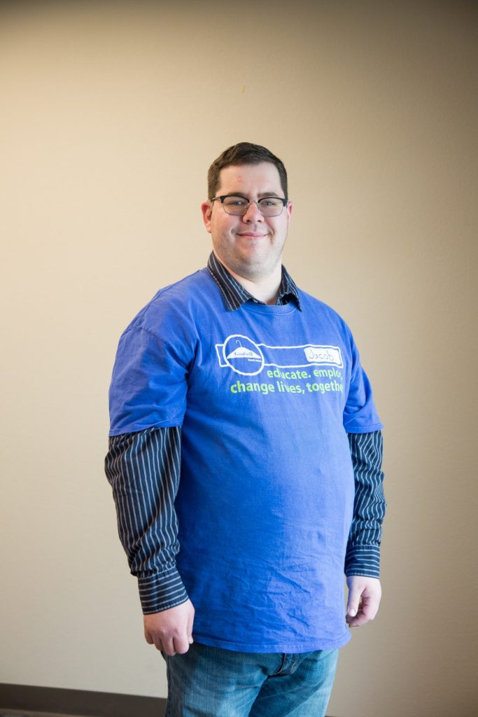 Smiling man wearing a blue T-shirt with a slogan, standing indoors against a beige wall.
