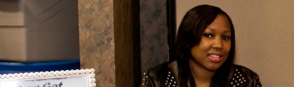 Young woman with long dark hair wearing a studded black jacket, smiling indoors.