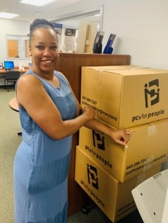 Smiling woman in a blue sleeveless dress stacks cardboard boxes labeled 'PCs for People' in an office.