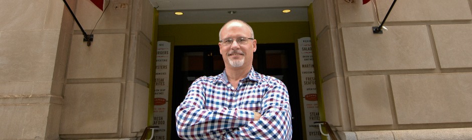 Man with glasses in a blue checkered shirt stands with arms crossed in the doorway of a restaurant.
