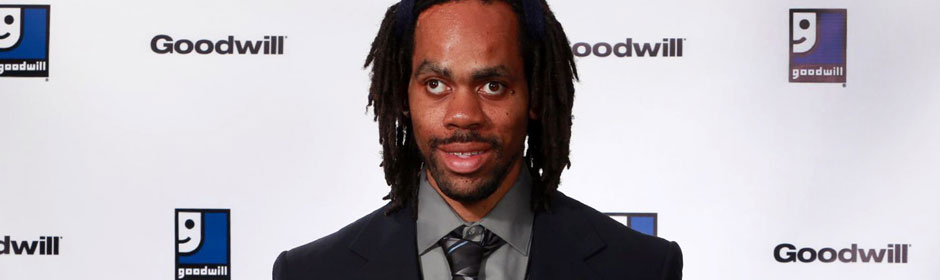 Man with dreadlocks wearing a suit at a Goodwill event in front of a branded backdrop.