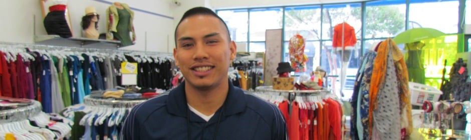Smiling man in a navy polo stands in a bright clothing store with racks of colorful shirts.
