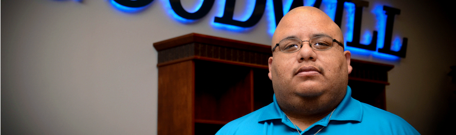 Bald man in a blue polo shirt stands in front of a wooden display with a blue illuminated sign in the background.