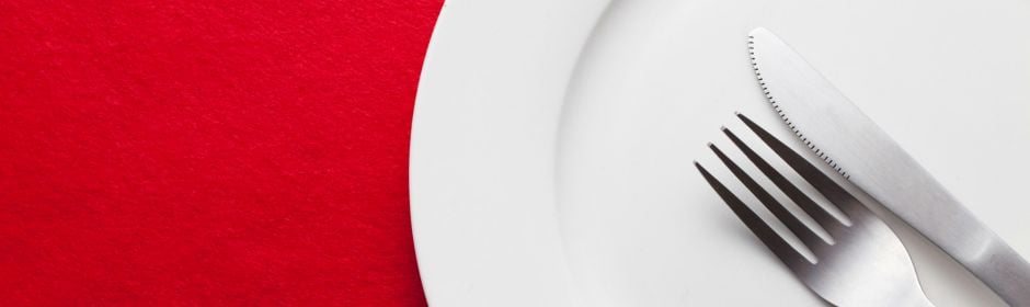 White plate with a knife and fork resting on the edge against a red background