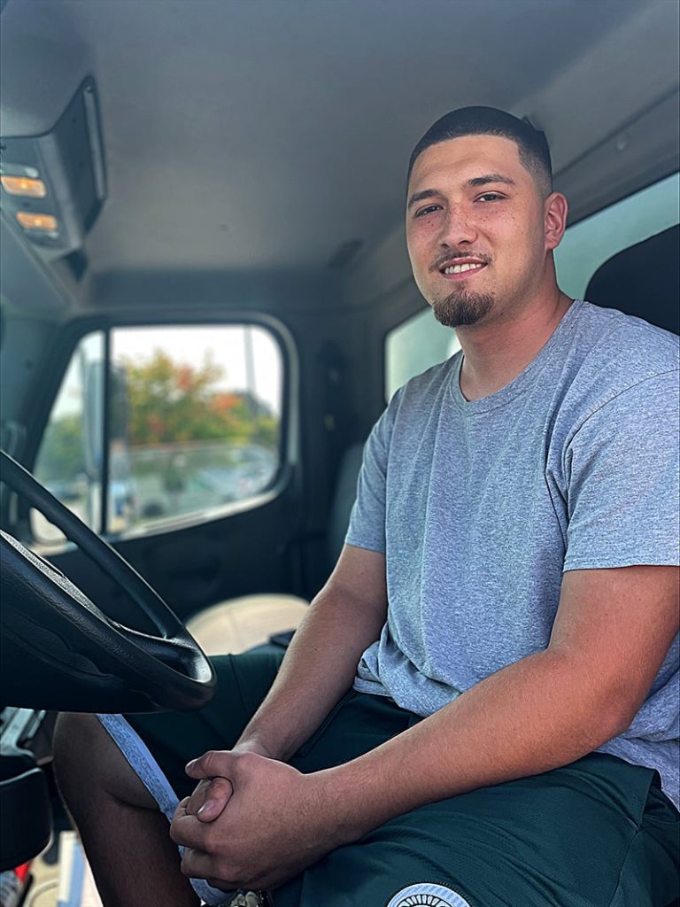 Male truck driver sits in the cab, smiling, with hands clasped in his lap.