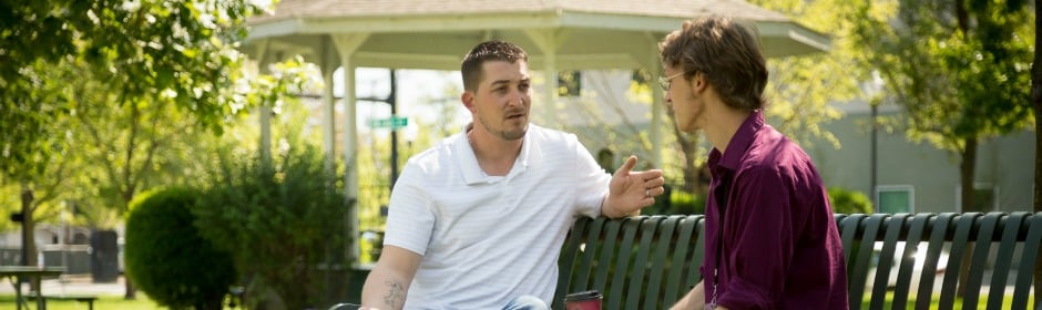 Two men chat on a sunny park bench with trees, greenery, and a gazebo in the background.