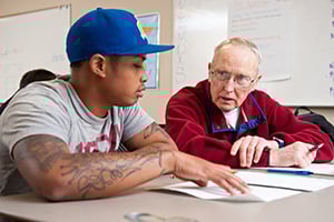 Young man with a blue cap and tattoos consults an older man with glasses in a classroom.