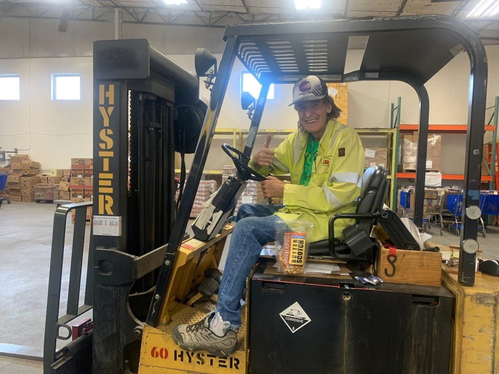 Worker in a yellow safety jacket sits on a forklift in a warehouse, giving a thumbs-up.