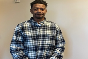 Man in blue and white plaid shirt standing indoors against a pale wall.